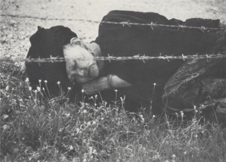 Old woman refugee in traditional dress sleeps curled up on he ground, facing outward, next to barbed wire fence of camp.