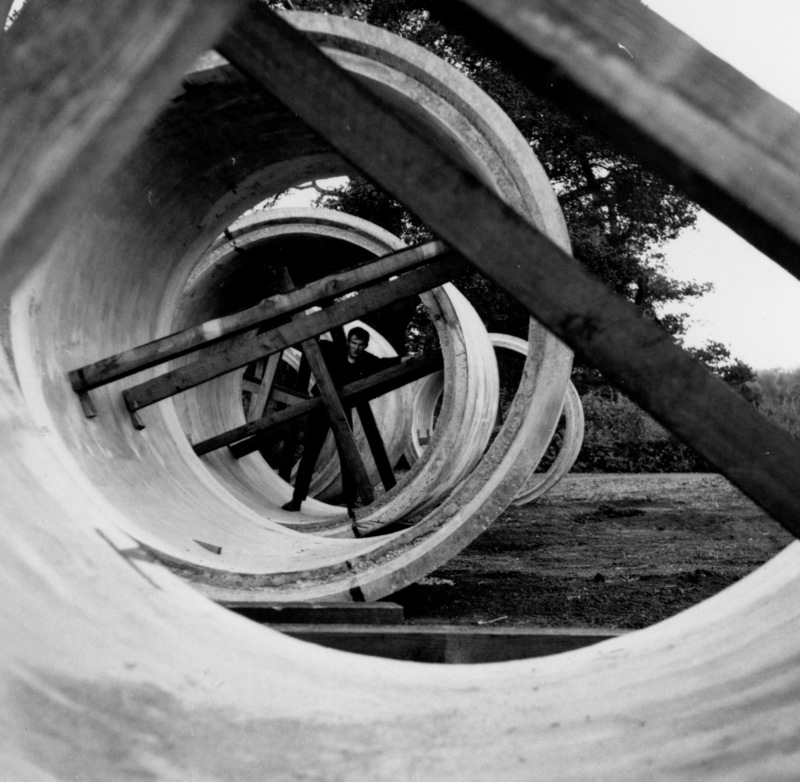 Large concrete sewage lines are set on the ground before being buried and connected. An unidentified man stands inside one of the concrete pipes, showing the massive size of these lines.