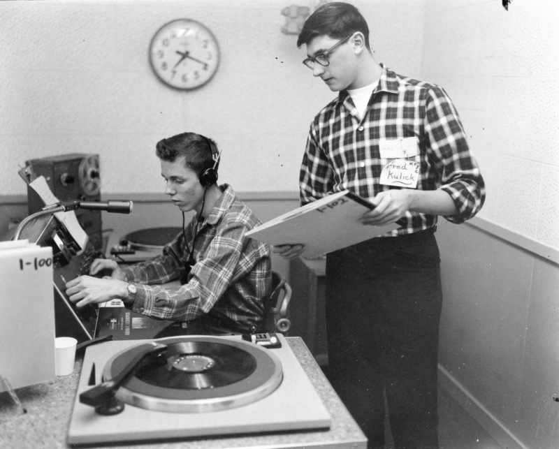 KFJC station manager Fred Kulick standing next to seated student at broadcast desk in the basement of Foothill College in Mountain View.