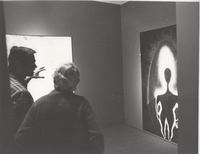 Two gallery viewers look at a painting with a dark body silhouette.

