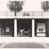Modernist concrete building, poster in one of three window bays, palm trees behind.