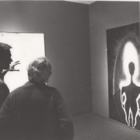 Two gallery viewers look at a painting with a dark body silhouette.
