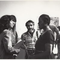 Two women in foreground talk. Male reporter listens. Video camera off to right.