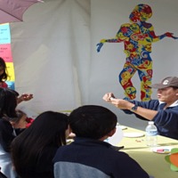 Male college student demonstrating art  to students around an art table.