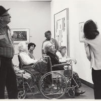Docent gives exhibition tour to seniors, several using wheelchairs.