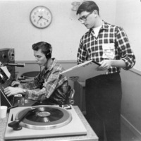 KFJC station manager Fred Kulick standing next to seated student at broadcast desk in the basement of Foothill College in Mountain View.